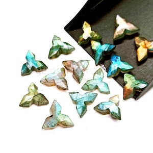 May include: A collection of faceted, tri-lobed gemstone beads in shades of green, blue, and gold. The beads are arranged on a white surface, with some spilling out of a black tray. The gemstones have a geometric design.