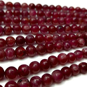 May include: A strand of deep red ruby beads, each bead is round and smooth with a slight shine. The beads are arranged in a loose pile, showing the individual beads.