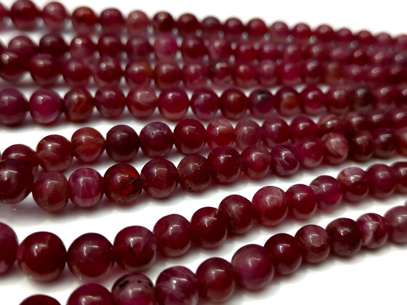 May include: A strand of deep red ruby beads, each bead is round and smooth with a slight shine. The beads are arranged in a loose pile, showing the individual beads.