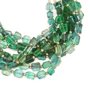 Peut inclure: Collier à plusieurs rangs avec des perles rectangulaires et facettées dans des tons de vert et de sarcelle. Les perles sont enfilées avec des accents métalliques argentés, créant un effet superposé. Le collier est sur fond blanc.
