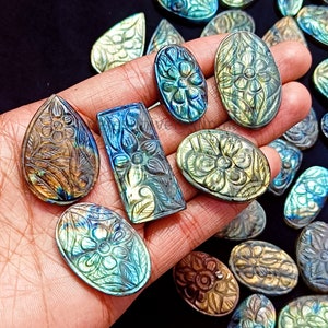 May include: A collection of labradorite gemstone cabochons with intricate carved floral designs. The cabochons are various shades of blue, green, and brown with iridescent flashes.
