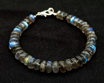 May include: A beaded bracelet featuring iridescent labradorite stones. The bracelet has a silver clasp and is composed of round, polished beads in shades of gray, blue, and brown. The bracelet is displayed against a black background.