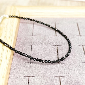 May include: A black beaded necklace with a silver clasp. The necklace is made of small, round beads and is lying on a textured surface.