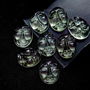 May include: Eight green face carvings, each with a unique expression, are arranged on a black surface. The carvings are oval-shaped and have a smooth, polished finish.