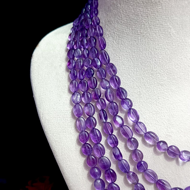 May include: A multi-strand necklace featuring oval-shaped amethyst beads in varying shades of purple. The beads are strung together, creating a cascading effect. The necklace is displayed on a white bust, with a dark background.