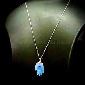 May include: A silver chain necklace with a blue hand pendant featuring a small red gemstone in the center. The pendant is a hamsa hand, a symbol of protection and good luck.