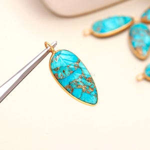 Könnte beinhalten: Ein tropfenförmiger Türkis-Anhänger mit einem goldfarbenen Metallrand und einem kleinen Verbindungsring an der Oberseite. Der Stein zeigt eine leuchtende blaue Farbe mit komplexer kupferfarbener Äderung auf der Oberfläche, gehalten von einer Pinzette.