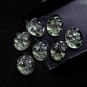 May include: Eight green faceted gemstone cabochons carved with a human face. The cabochons are oval shaped and have a smooth, polished surface. The cabochons are arranged in a semi-circle on a black surface. The text "gemsuniverseindia" is visible in the top right corner of the image.