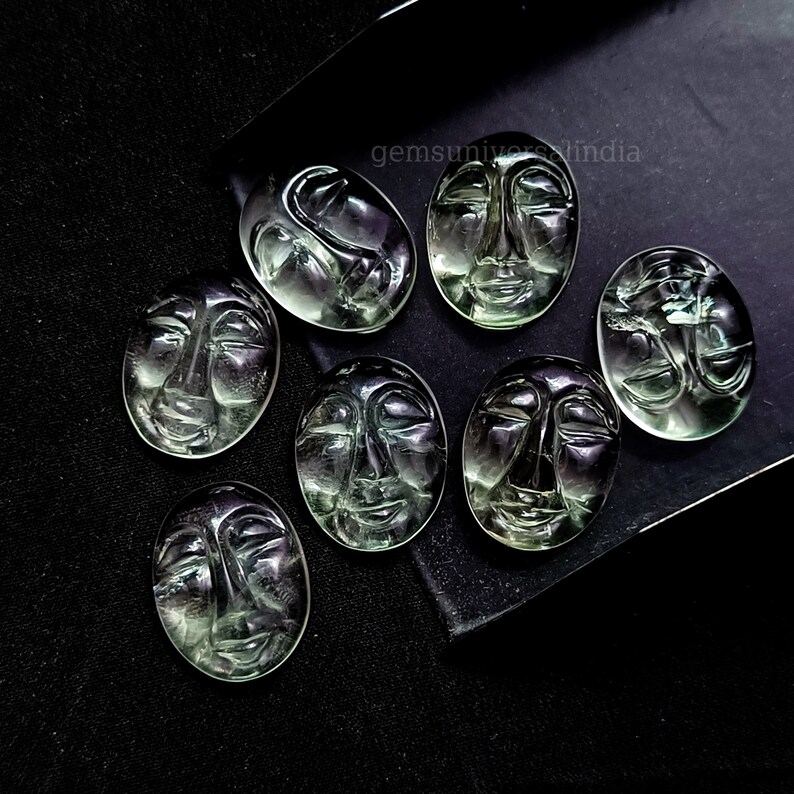 May include: Eight green faceted gemstone cabochons carved with a human face. The cabochons are oval shaped and have a smooth, polished surface. The cabochons are arranged in a semi-circle on a black surface. The text "gemsuniverseindia" is visible in the top right corner of the image.