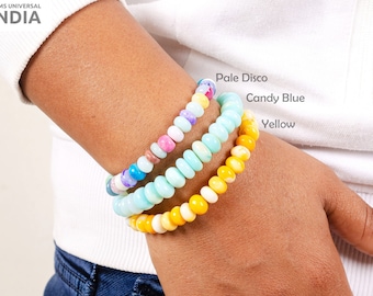 May include: Three colorful beaded bracelets stacked on a wrist. The top bracelet is pale blue with white swirls, the middle bracelet is candy blue with pink and white beads, and the bottom bracelet is yellow.