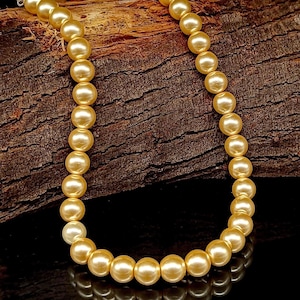 May include: A strand of golden-colored faux pearls. The pearls are round and evenly sized, strung together to form a necklace. The necklace is displayed against a dark, textured wooden background, creating a contrast with the luminous pearls.