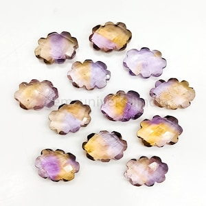 Pode incluir: Uma coleção de pedras preciosas facetadas em forma de nuvem. As pedras exibem um gradiente de cores, incluindo tons de roxo, amarelo e castanho. As pedras preciosas estão dispostas sobre uma superfície branca, mostrando suas formas e variações de cores únicas.