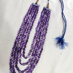 May include: A multi-strand necklace featuring oval-shaped, deep purple amethyst beads. The necklace has a blue cord with a tassel and decorative accents at the top. The beads are strung together in multiple layers.