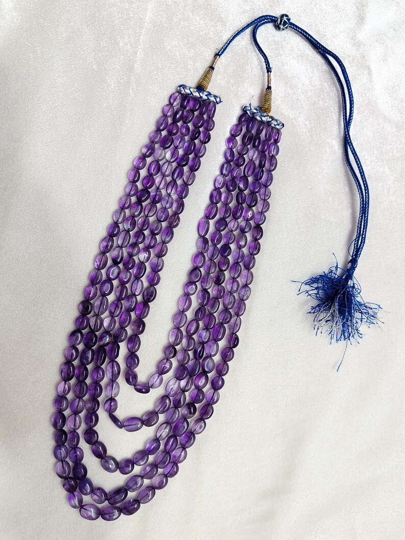 May include: A multi-strand necklace featuring oval-shaped, deep purple amethyst beads. The necklace has a blue cord with a tassel and decorative accents at the top. The beads are strung together in multiple layers.