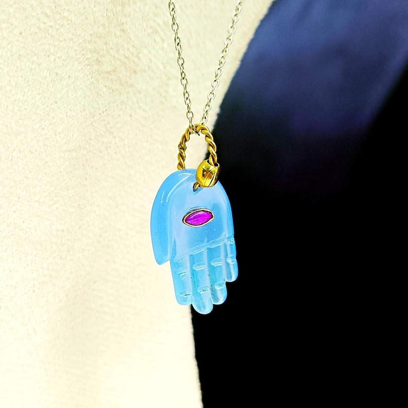 May include: A light blue hand-shaped pendant with a pink gemstone eye, hanging from a gold chain.