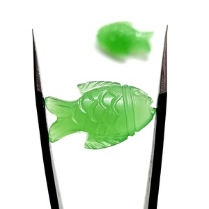 K&ouml;nnte beinhalten: Zwei gr&uuml;ne Jade-Fischschnitzereien, eine mit einer Pinzette gehalten, die andere auf wei&szlig;em Hintergrund.