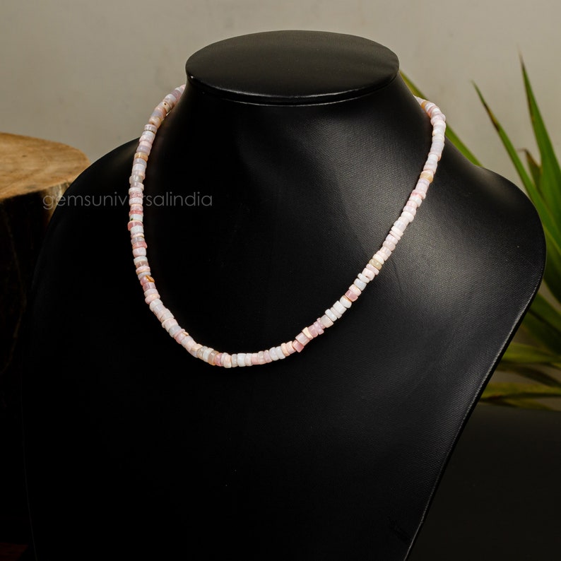 May include: A pink and white beaded necklace made of small, square-shaped beads. The necklace is displayed on a black mannequin.