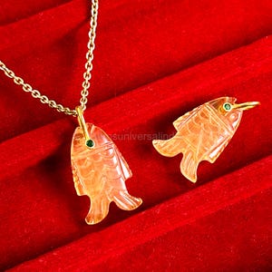 May include: A gold-toned necklace with a fish pendant and a matching fish-shaped earring. The fish pendants are carved from a translucent orange material and feature a small green gem. The necklace and earring are displayed on a red surface.