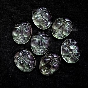 May include: Seven green and purple faceted gemstone cabochons carved with different facial expressions. The cabochons are oval shaped and have a smooth, polished finish.