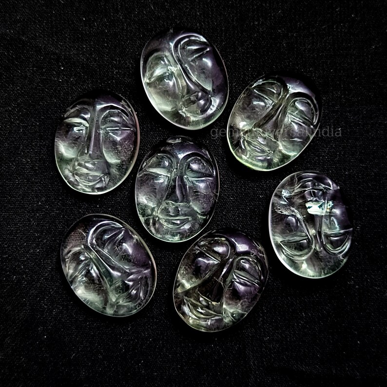 May include: Seven green and purple faceted gemstone cabochons carved with different facial expressions. The cabochons are oval shaped and have a smooth, polished finish.