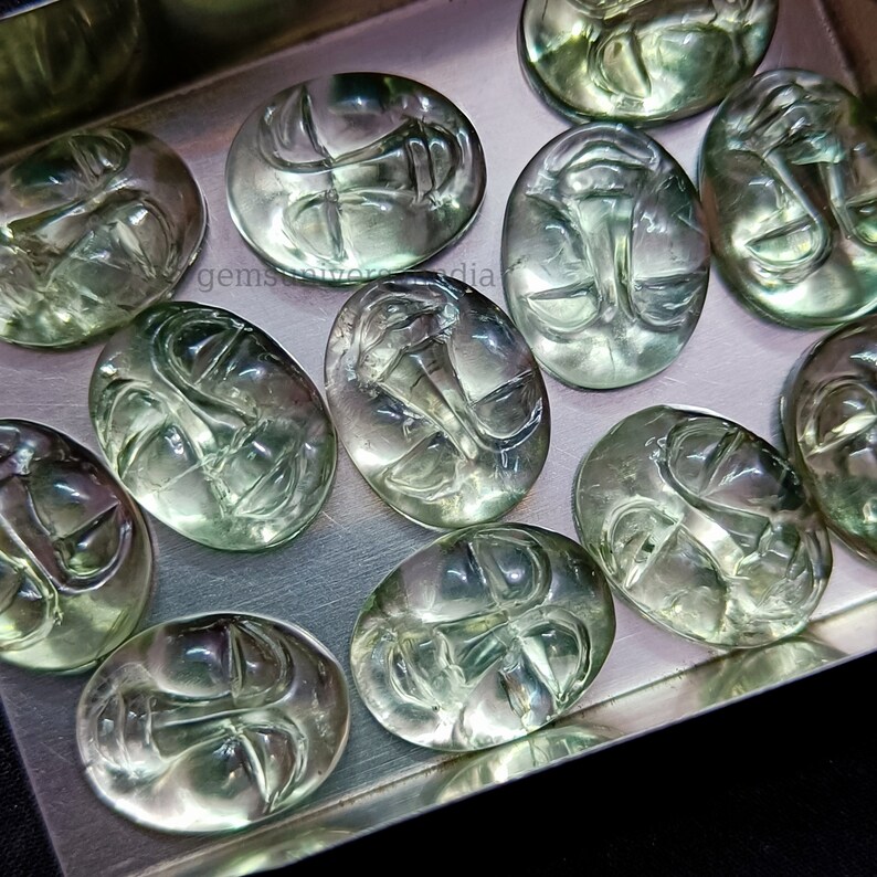May include: A collection of green faceted gemstone cabochons with carved faces. The cabochons are oval shaped and have a translucent appearance.