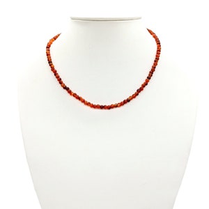 Puede incluir: Un collar hecho de peque&ntilde;as cuentas redondas de cornalina naranja. El collar est&aacute; en un busto de maniqu&iacute; blanco.