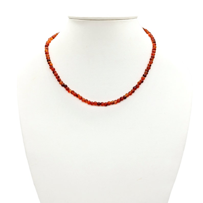 Puede incluir: Un collar hecho de peque&ntilde;as cuentas redondas de cornalina naranja. El collar est&aacute; en un busto de maniqu&iacute; blanco.