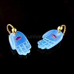 May include: A pair of blue glass earrings shaped like hands with a small red gemstone in the center of each hand. The earrings are attached to gold hoops.