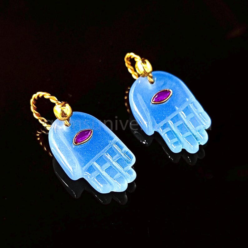 May include: A pair of blue hamsa hand earrings with a purple gemstone in the center of each hand. The earrings are made of gold and have a delicate design.