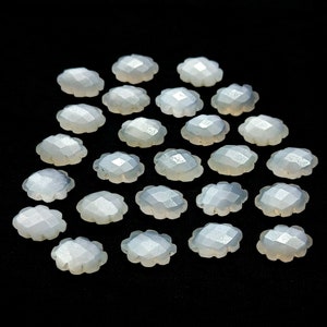 May include: A collection of 20 white, faceted, gemstone cabochons in a flower shape. The cabochons are arranged in a semi-circle on a black background.