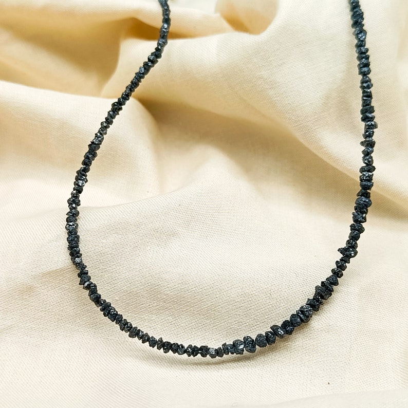 May include: A black diamond necklace made of small, rough-cut stones. The necklace is strung on a thin, silver chain and is displayed on a cream-colored fabric background.