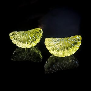 May include: Two carved, translucent, yellow-green gemstone slices, possibly citrine, with intricate leaf-like patterns. The gemstones are displayed on a reflective black surface, creating mirrored reflections. The overall aesthetic is elegant and natural.