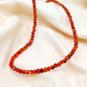 Puede incluir: Un collar hecho de peque&ntilde;as cuentas redondas de cornalina naranja. Las cuentas est&aacute;n enhebradas en un cord&oacute;n delgado y el collar est&aacute; sobre un fondo de tela blanca.