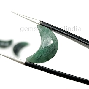 May include: A green gemstone carved into a crescent moon shape. The gemstone is held by tweezers against a white background.