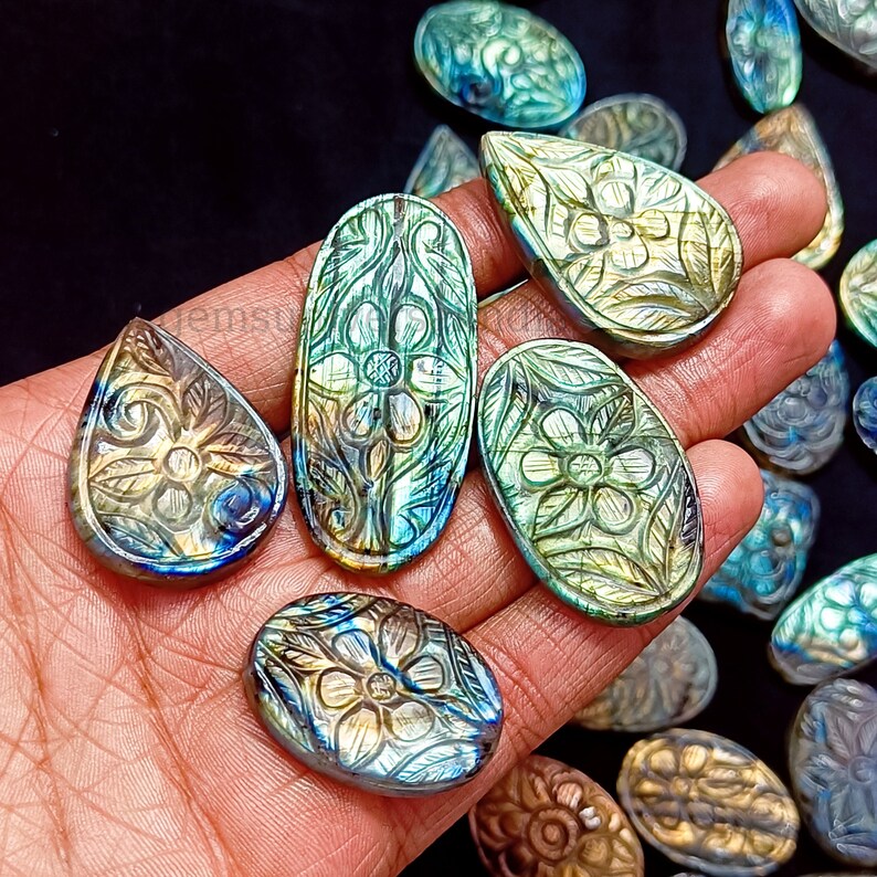May include: A collection of carved labradorite gemstone cabochons with intricate floral designs. The stones are a mix of green, blue, and gold colors with iridescent flashes.