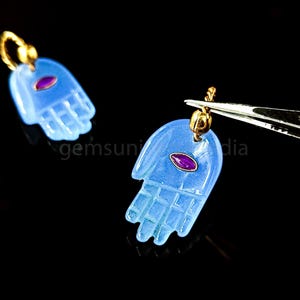 May include: Two blue hamsa hand charms with gold accents and a red gemstone in the center. The charms are hanging from gold loops.