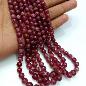 May include: A strand of deep red ruby beads, each bead is round and smooth with a subtle striated pattern. The beads are strung together on a clear thread.