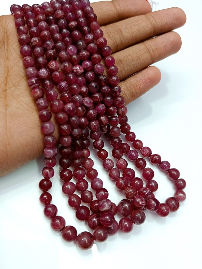 May include: A strand of deep red ruby beads, each bead is round and smooth with a subtle striated pattern. The beads are strung together on a clear thread.