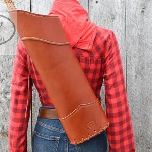 May include: A tan leather quiver with visible stitching along the edges and a reinforced base. The quiver features a smooth finish and a structured design, with a small embossed logo near the bottom edge, worn over a red and black plaid shirt.