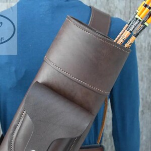 May include: A dark brown leather quiver with a structured cylindrical body, a front pocket, and visible stitching. The quiver is worn over the shoulder and contains several arrows with orange and black fletching. The logo gw leathercraft is visible.
