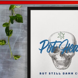May include: A black framed print with a white background. The print features a black and white illustration of a skull with a marijuana leaf on its forehead. The text "Pot Head" is written in blue cursive above the skull. The text "But Still Damn Cool" is written in blue below the skull.