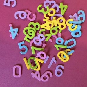 Könnte beinhalten: Eine Ansammlung bunter Schaumstoffzahlen in verschiedenen Größen und Farben, darunter Rosa, Grün, Gelb und Blau, auf einem magentafarbenen Hintergrund. Die Zahlen von Null bis Zehn eignen sich für Bildungszwecke.