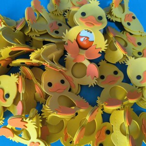 May include: A group of yellow foam rubber ducklings with orange beaks and feet. The ducklings are arranged in a pile, with some of them overlapping. The ducklings have a hole in the center of their bodies, which is meant to hold an Easter egg.