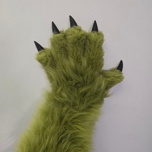 Op de afbeelding: Een groene, harige hand met zwarte klauwen. De hand is omhoog geheven met de vingers gespreid.
