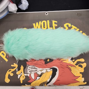 Op de afbeelding: Een lichtgroene, pluche staart met een zilveren clip. De staart ligt op een zwart oppervlak met de tekst "Wolf Studios" in gele letters.