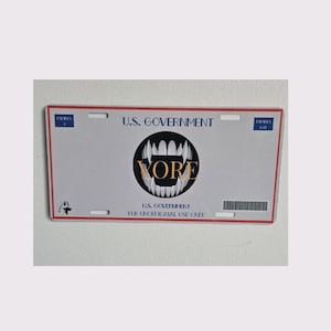 May include: A rectangular license plate with a white background and a red border. The text "U.S. GOVERNMENT" is at the top, and a black circle with a toothy design and the word "VORE" is in the center. The bottom text reads "U.S. GOVERNMENT FOR UNOFFICIAL USE ONLY."