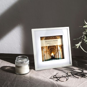 May include: White square picture frame with a 3D effect. The frame is 15.24 cm x 15.24 cm and has a white mat. The frame is sitting on a table with a candle and glasses.