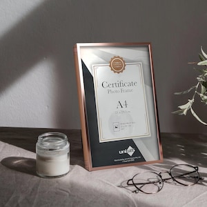 May include: A rose gold A4 certificate photo frame with a black and white interior. The frame has a white label with the text "Certificate Photo Frame A4 21 x 29.7cm" and a smaller label with the text "Inside Print Size: 21 x 29.7cm A4". The frame is made by Unibift.