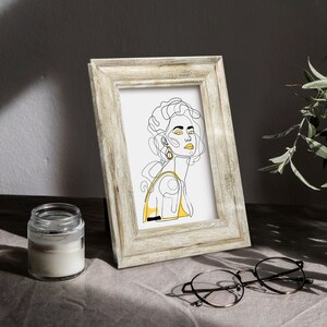 May include: A framed line drawing of a woman with yellow accents. The drawing is in a light wood frame and is sitting on a table with a candle and glasses.