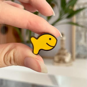 May include: A yellow enamel pin in the shape of a smiling goldfish cracker. The pin has a black outline and a small black dot for the eye. The pin is held between two fingers.
